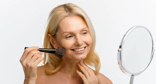 Sophisticated blonde 50s woman looking and her reflection in mirror and dioung makeup, beautiful middle-aged lady enjoying effect of skin care treatment, holding brush near face with smooth skin