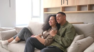 Side view of the multiracial couple chilling on the couch, watching movie in the studio apartment. Guy switching channel with TV remote