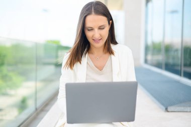 Carefree female office employee typing on the laptop sitting on the step in the city, businesswoman or freelancer woman responds to emails, working outdoors, urban landscape on the background