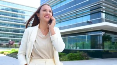 Businesswoman in smart casual white suit having phone talk walking in the city during break, smiling female employee speaking on smartphone outdoors with skyscrapers on the background