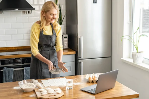 Blonde middle-aged woman cooks at home in the kitchen, housewife in apron watches a video recipe on a laptop and works with the dough on the table