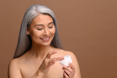 Yakışıklı, orta yaşlı, çıplak omuzlu bir Asyalı kadın elinde izole edilmiş bej arka planında yaşlanma önleyici bir kavanoz dolusu nemlendiriciye bakıyor. Spa ve rahatlama, kozmetoloji prosedürü
