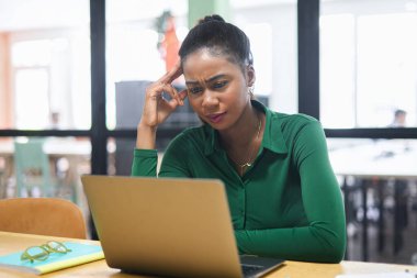 Thoughtful african american female office employee using laptop indoors, looking at laptop screen with puzzled face expression, businesswoman solving tasks