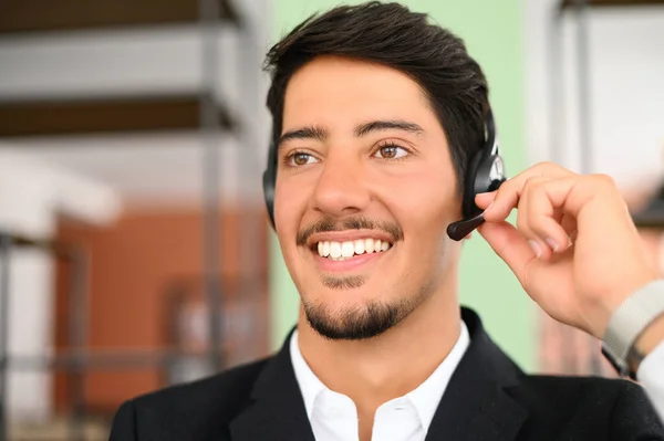 Smiling young latin male office employee wearing using headset and looking aside, positive man with the beard working in customer service department, making and receiving calls, sitting on workplace