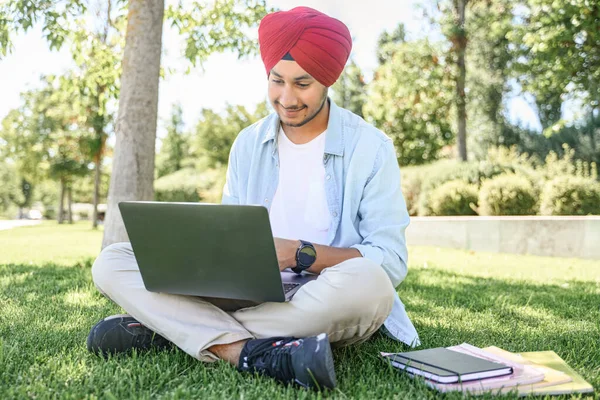 Serene Indian male student in traditional turban using laptop for online studying sitting outdoors on the lawn in campus area, preparing homework, researching topic, positive hindu guy typing