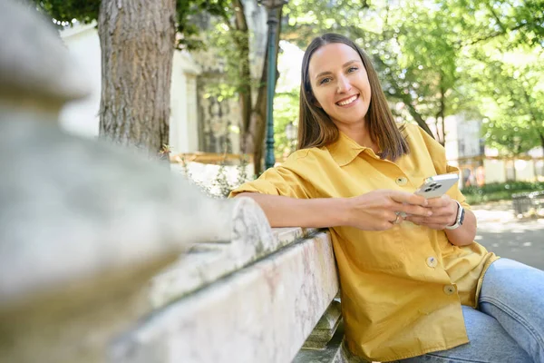 Attractive cheerful woman using smartphone sitting in the park, smiling female student in yellow casual shirt holding phone and looking at the camera, texting online, usingnew mobile app