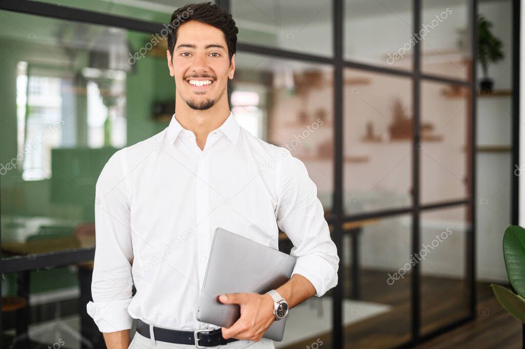 Working with joy. Smiling cheerful hispanic business man wearing formal white shirt standing with laptop computer in office, looking at camera, proud and ambitious latin start-up owner