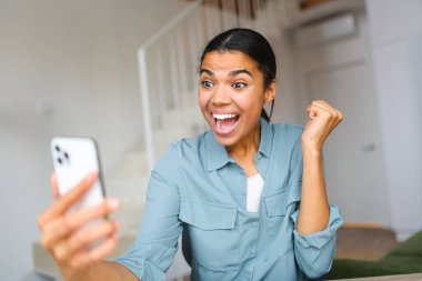 Kanepede otururken ve elinde akıllı telefon tutarken rahat giyinen heyecanlı genç bir kadın. İnternetteki kadın piyangoda kazandı, en iyi anlaşmayı kutladı, zafer işareti olarak yumruğunu sıktı.