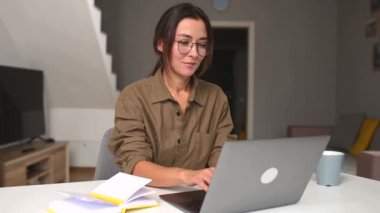 Concentrated young Asian businesswoman in casual wear and glasses sitting at the desk in the home office, working remotely on the laptop. Smiling freelancer woman typing, answering emails