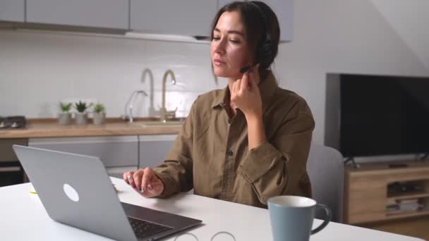 若い陽気な女性は 自宅のオフィスでデスクに座ってオンラインで話すためにヘッドセットを使用しています 画面を見ている快適な女性オペレーター リモートワークの概念 — ストック動画