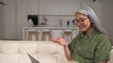 Lovely elderly woman smiling, chatting with family, using a laptop for video connection, remote meeting, say hi, glad to see, lying on the couch. Video call concept
