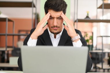 Worried hispanic man sits at the desk in front of laptop in modern office, looks at the screen and holds temples, concerned about solving tasks, male office employee full of doubt