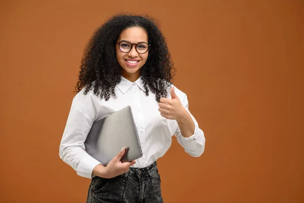Young African American woman with afro hairstyle wearing smart casual wear standing isolated on brown and showing big finger, carrying laptop computer, smiling, female office employee, student