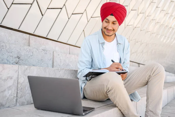 Indian student man in traditional turban sitting outdoors and using laptop for learning on the distance, hindu guy watching webinar and taking notes, male student studying online, e-learning