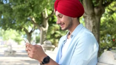 Indian male freelancer, student, entrepreneur wearing red national turban using smartphone sitting in the park outdoors, guy with phone in traditional headwrap texting, chatting online