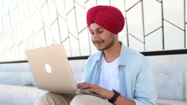 Indian freelancer man in traditional turban sitting outdoors with laptop, developer working on the distance, young guy enjoys remote work, male student studying online, e-learning concept