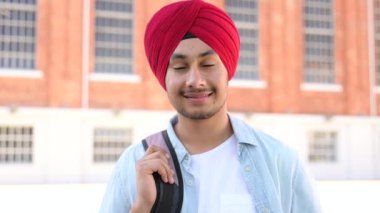 Inspired Indian male student in traditional turban with backpack outdoors, foreigner guy proud to be university member, looking at the camera standing outdoors