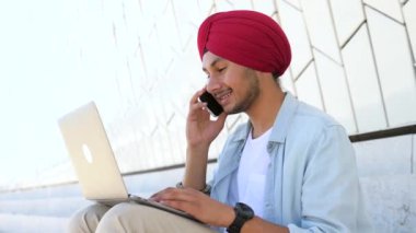 Cheerful Indian male entrepreneur in traditional headwrap and casual shirt sitting on the steps in city with laptop and talking on the smartphone, making schedule