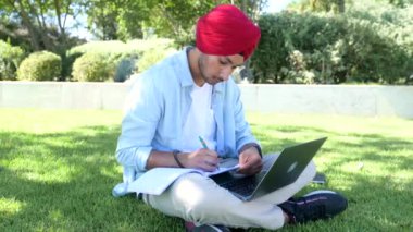 Geleneksel türbanlı sakin Hintli erkek öğrenci çevrimiçi çalışmak için dizüstü bilgisayar kullanıyor kampüs bölgesinde çimlerin üzerinde oturuyor, ödev hazırlıyor, konu araştırıyor, pozitif Hintli adam yazı yazıyor.