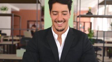 Confident latin businessman using laptop in the office. Young male office employee in formal wear typing and chatting online, develops software indoors, looks at computer screen with smile