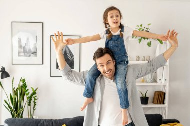Kollarını açan tatlı kız uçak numarası yapıyor ve gülümsüyor. Sevgi dolu bir baba, küçük kızını omuzlarında taşıyarak rahat bir oturma odasında kanepede oturuyor, evde çocukla oynayan bekar bir ebeveyn.
