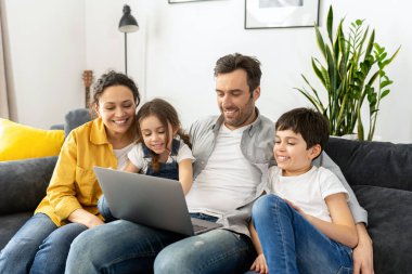Family of four using laptop sitting on the sofa in cozy apartment, parents and two kids watching movie on computer, comedy, spends time on the weekend together at home
