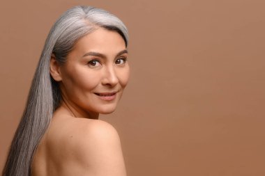 Close-up portrait of fresh and gorgeous middle-aged woman with pure and healthy skin and grey hair, stands with naked shoulders isolated on brown background, mature lady looks at camera and smiles