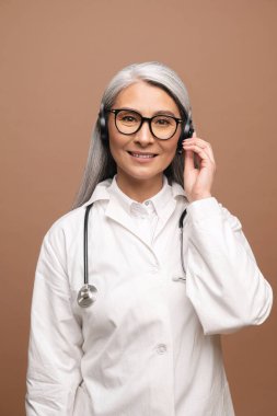 Senior Asian woman doctor in white medical gown with stethoscope using wireless headset for call to patient, looking at camera standing isolated on brown. Consulting and therapy online