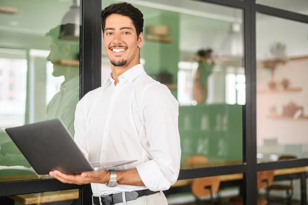 Portrait of young latin guy standing and holding laptop in contemporary office space, male white collar worker looks at camera, posing indoors with glass loft style partition on background