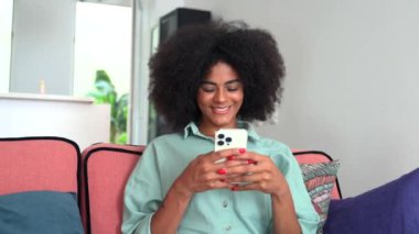 Young smiling african woman sitting at the sofa at home and using smartphone, texting, using smartphone for online shopping. Technologies and people concept