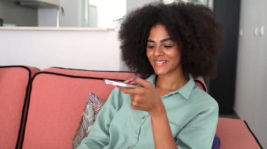 Smiling African-american woman with curly hair holding phone and speaking on speakerphone, multiracial girl recording and sending voice message, using voice typing feature on smartphone at home