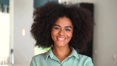 Headshot of a young African American ethnic female with Afro curly hairstyle and beautiful smile looking at the camera standing against blurred home interior on the background, satisfied and pleased