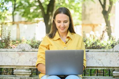 Optimistic young woman using laptop sitting on the bench in campus area, female student watching webinars, educational courses, learning on the distance, typing emails