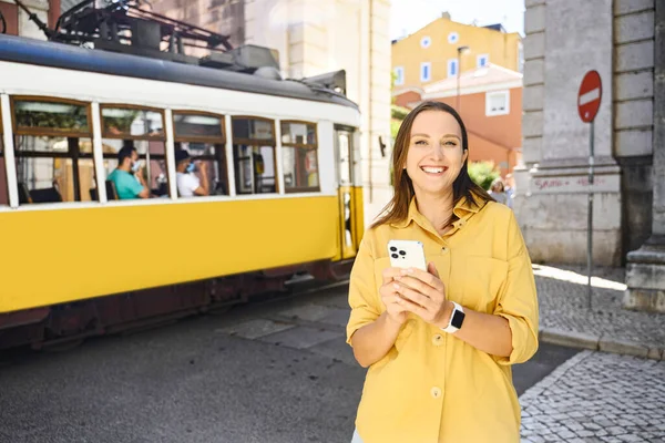 Smiling happy foreign woman tourist using smartphone with famous yellow tram on background, walking in old town of Lisbon, Portugal