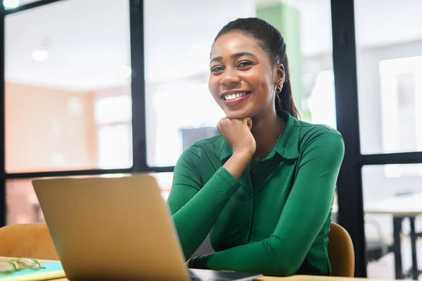 Happy inspired African-American female office employee using laptop indoors, resting chin with hand and looking at camera, smiles. Excited freelancer woman enjoys remote work in coworking space
