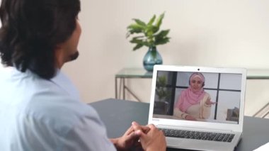 Indian male colleague talking online with Islamic female colleague, greeting each other during video meeting. Back view male student takes an online classes, woman in hijab on the screen