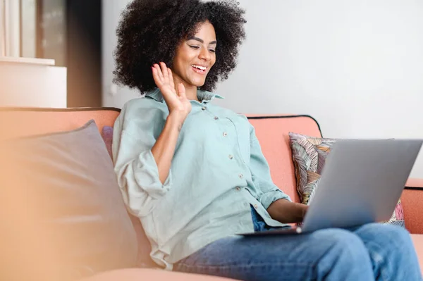 Video conference. Smiling young african american woman using laptop for making video call at home, waving at webcamera, sitting on the sofa