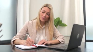 Concentrated businesswoman in smart casual wear using laptop for watching online webinar, takes a part in educational course or conference, female employee sitting at the desk in office, taking notes