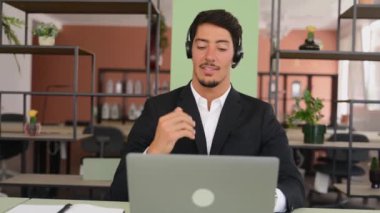 Front view at smiling latin male employee wearing headset and smart casual wear using laptop for connection with colleagues or customers, talking online sitting near windows in the office