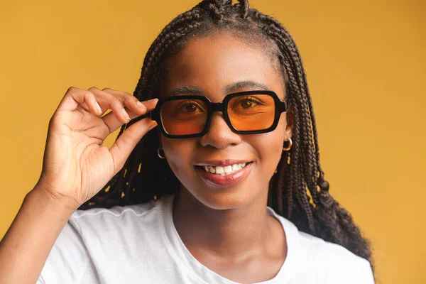 Beautiful positive african american lady smiles isolated on yellow background. Young brunette woman wearing sunglasses stands and looks at the camera