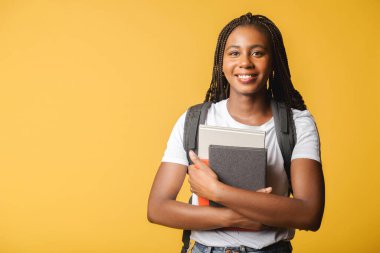 Happy excited female student with backpack standing isolated on yellow and holding books and abstracts, ready to studying, back to school concept