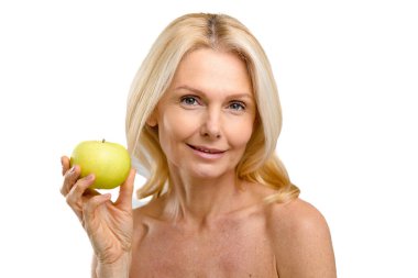 Cheerful joyful mature 50s woman posing with fresh green apple and smiling. Middle-aged model with naked shoulders isolated on white, demonstrates smooth and radiant skin. Anti age treatment