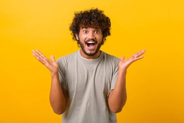 Astonished Indian young curly man screams wow and looking at the camera with mouth wide opened, surprised happy guy excited with incredible promotion, standing isolated on yellow