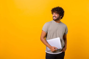 Waist up portrait of the smiling male person holding laptop in hands and smiling, Indian freelancer man standing isolated on yellow background, copy space