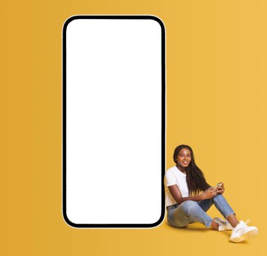 Smiling attractive girl sitting near huge smartphone with empty blank screen, looking at the camera and smiles, hipster woman advertising new app or website, mockup