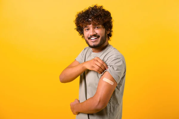 Smiling young curly Indian guy showing arm with band-aid after vaccine injection isolated on yellow background, vaccinated multiracial man feels safe with vaccine