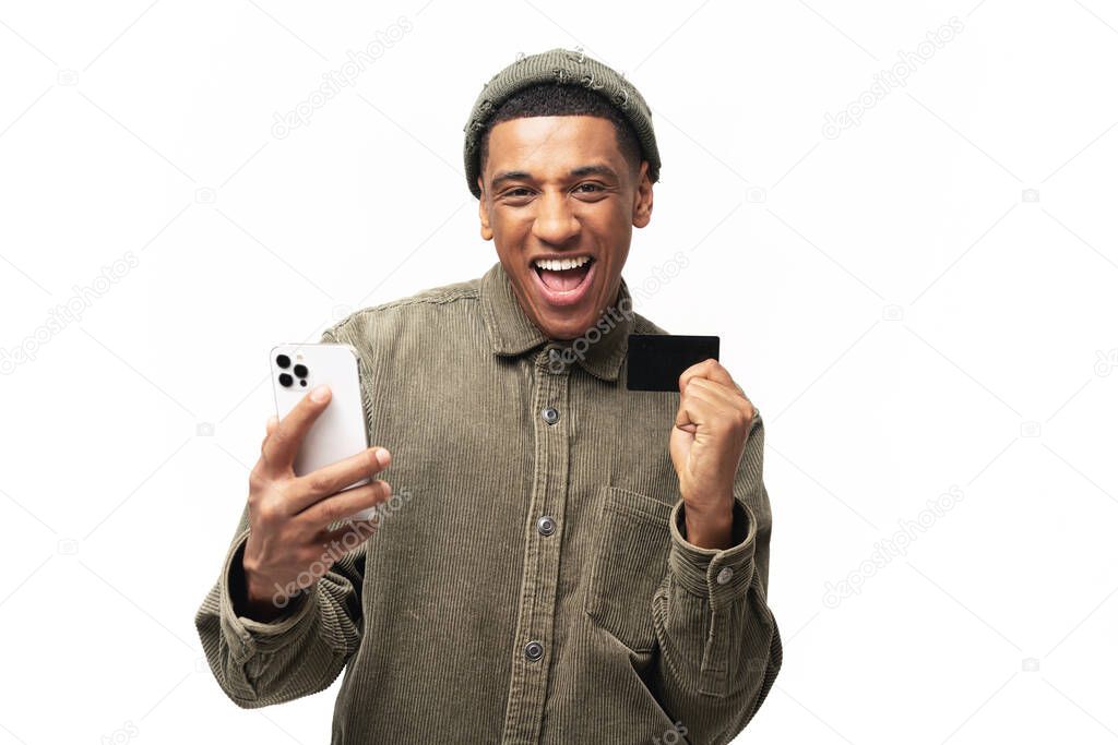 Blissful surprised young multiracial man holding debit card and smartphone and looks at camera with mouth open, isolated. Lucky man in casual clothes receive cash back from online purchases