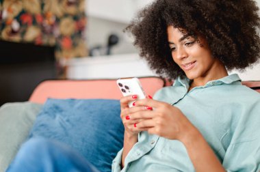 Side view at smiling calm young black woman with afro hairstyle sitting in relaxed pose on the sofa at home and using smartphone, texting, replying emails, typing sms, playing mobile game
