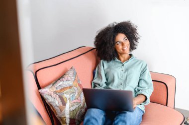 Optimistic african-american woman in casual wear using laptop while sitting on the couch. Young female student watching webinars, educational courses, learning on the distance, typing emails