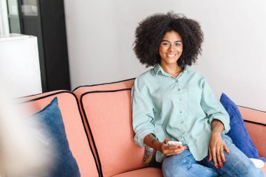 Portrait of beautiful black woman with natural curly afro hairstyle looking at the camera in cozy home atmosphere, charming serene and carefree girl holding smartphone and rest at home
